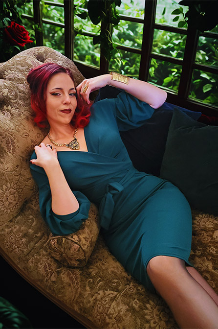 Author Devora Gray with red hair is lounging by garden in blue green dress